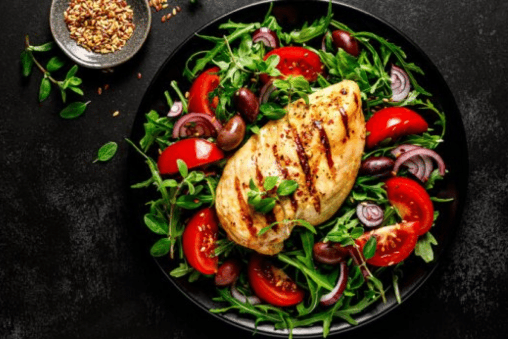 Chicken breast fillet grilled and fresh vegetable green salad with arugula, tomatoes and olives on black background, healthy food, mediterranean diet. Image source: St Adobe Stock