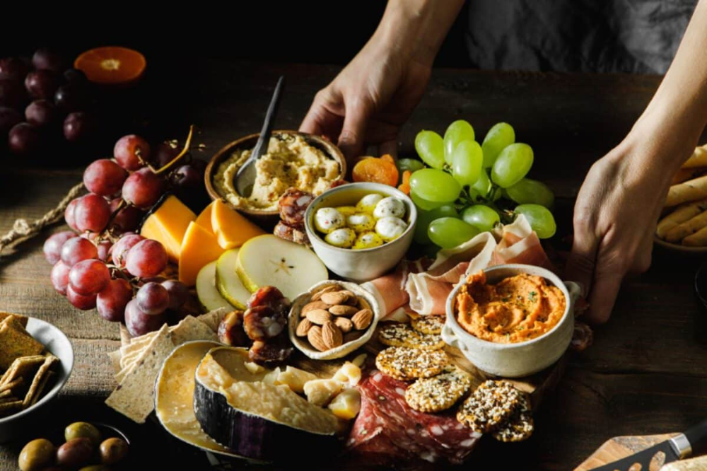 Close-up of a woman serving cheese and meat platter with fruits. Female hands placing mixed fruits with cheese and meat on wooden board. Image source: iStock